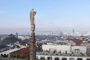 Blick vom Dach des Doms nach Norden auf eine der zahllosen Statuen auf einer Fiale. Links ist die Kuppel der Galleria Vittorio Emanuele II, unten die Dachterrassen und im Hintergrund die zahlreichen Hochäuser